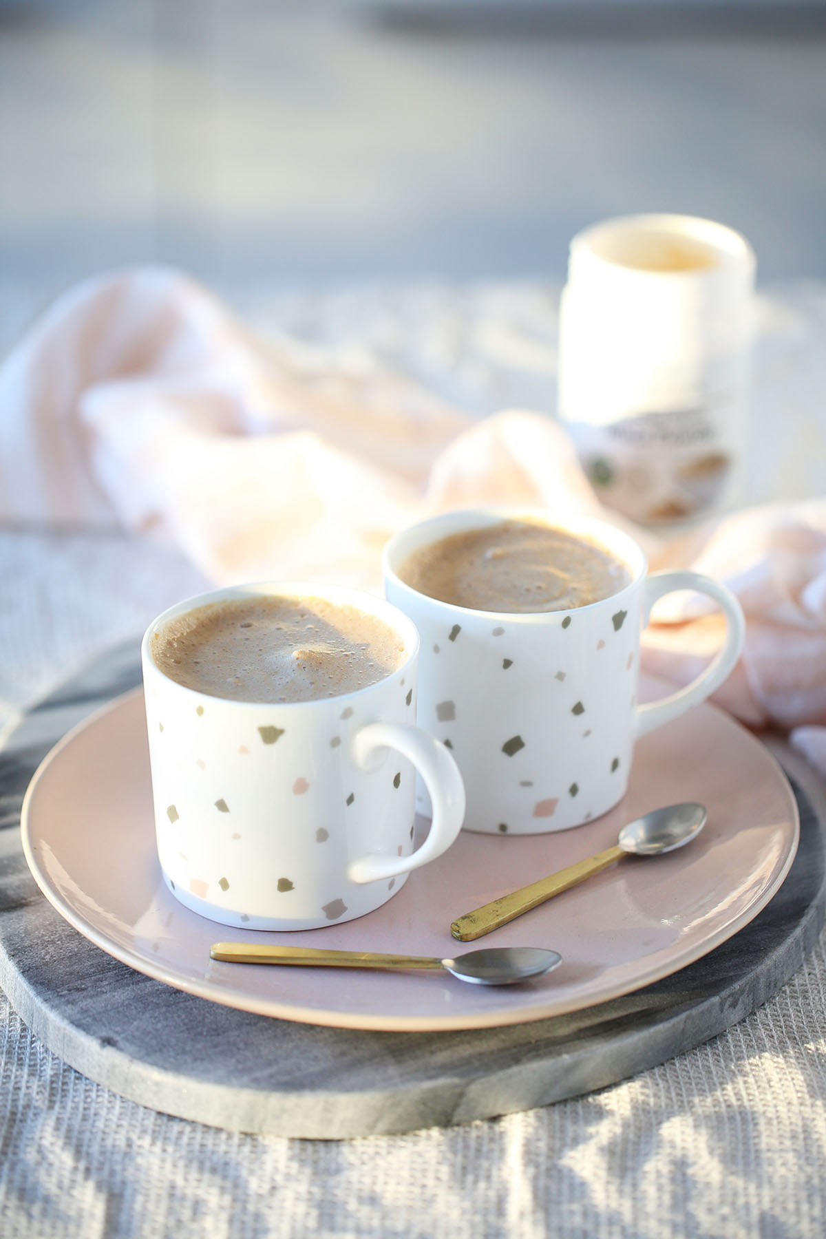 Two caramel maca lattes in spotty pastel mugs with gold spoons on a pink plate
