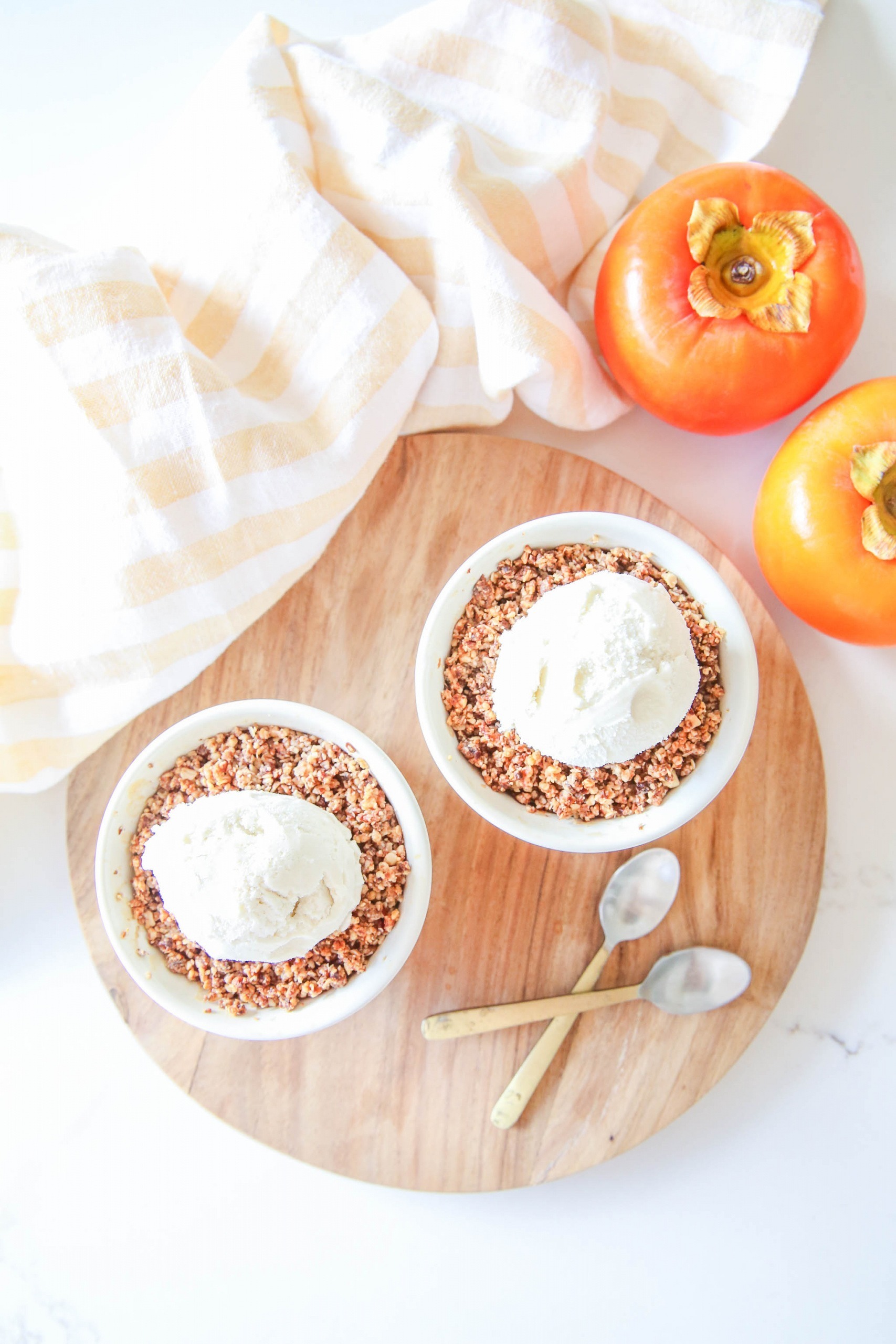 Two Persimmon and Apple Crumbles with a scoop of ice cream on top, placed on a chopping board. Two Persimmon placed in the corner