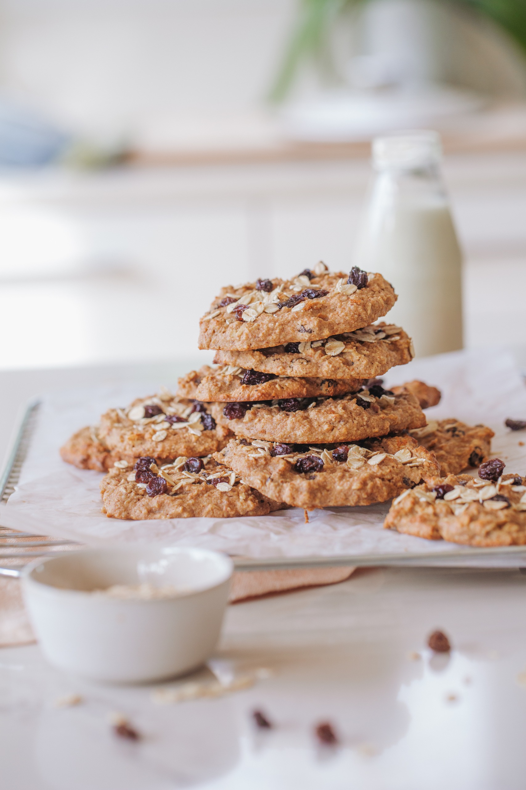 stacked Oat and Raisin Cookies on baking tray with white paper and bowl of oats and milk bottle