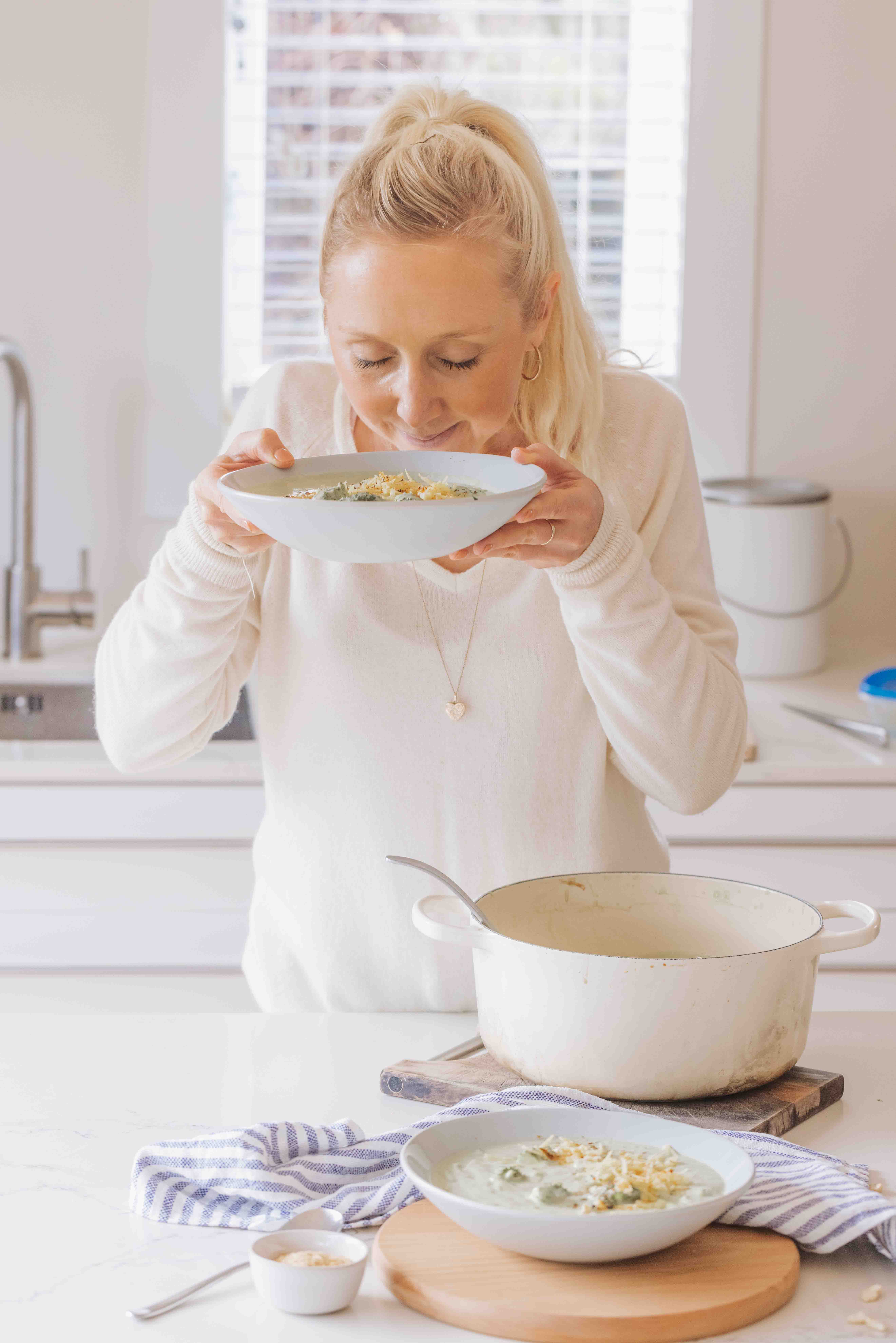Woman (Buffy Ellen Gill) smelling a bowl of creamy broccoli soup, with a large Le Creuset crock pot in front.