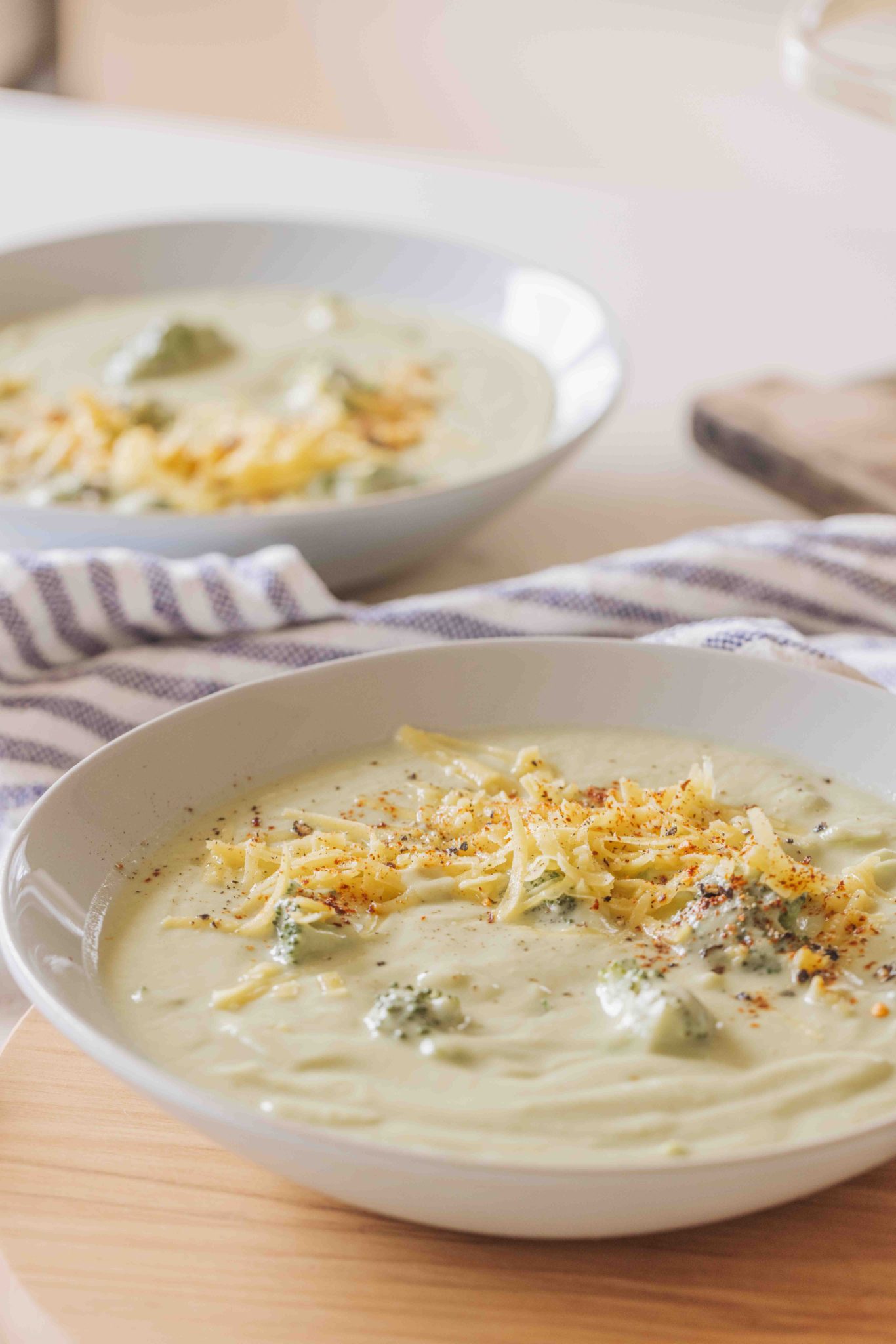 Two bowls of creamy broccoli soup, topped with grated cheese and red chilli flakes.