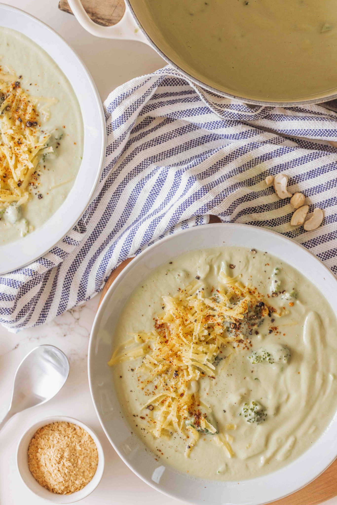 Two bowls of creamy broccoli soup, topped with grated cheese and red chilli flakes, with cashews on the side.