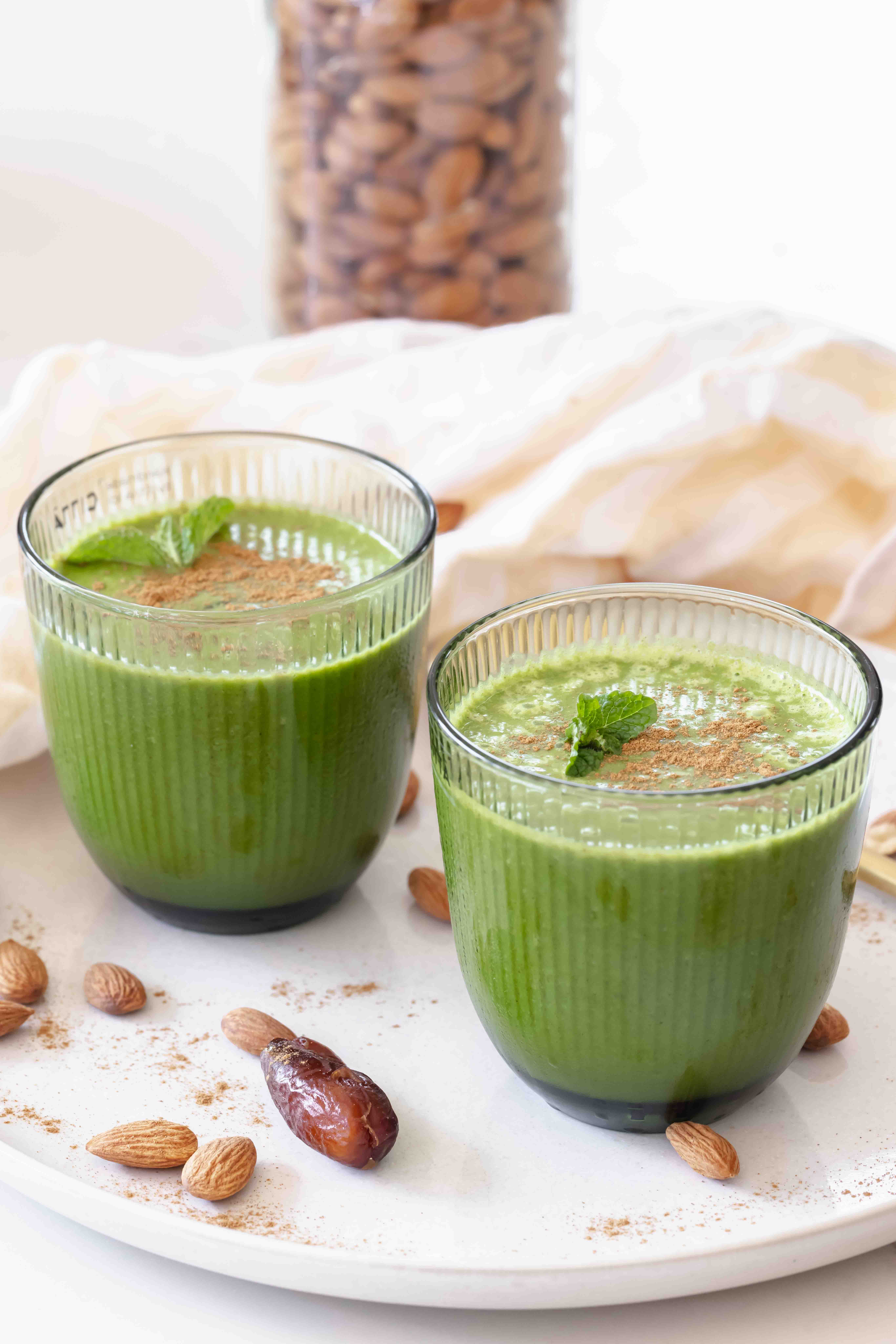 clear glass of green juice on white plate with dates, almonds, mint, and cinnamon