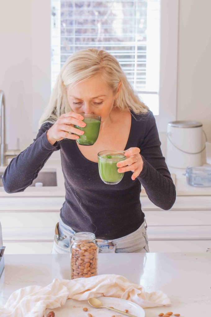 buffy ellen drinking clear glass of green juice on white plate with dates, almonds, mint, and cinnamon