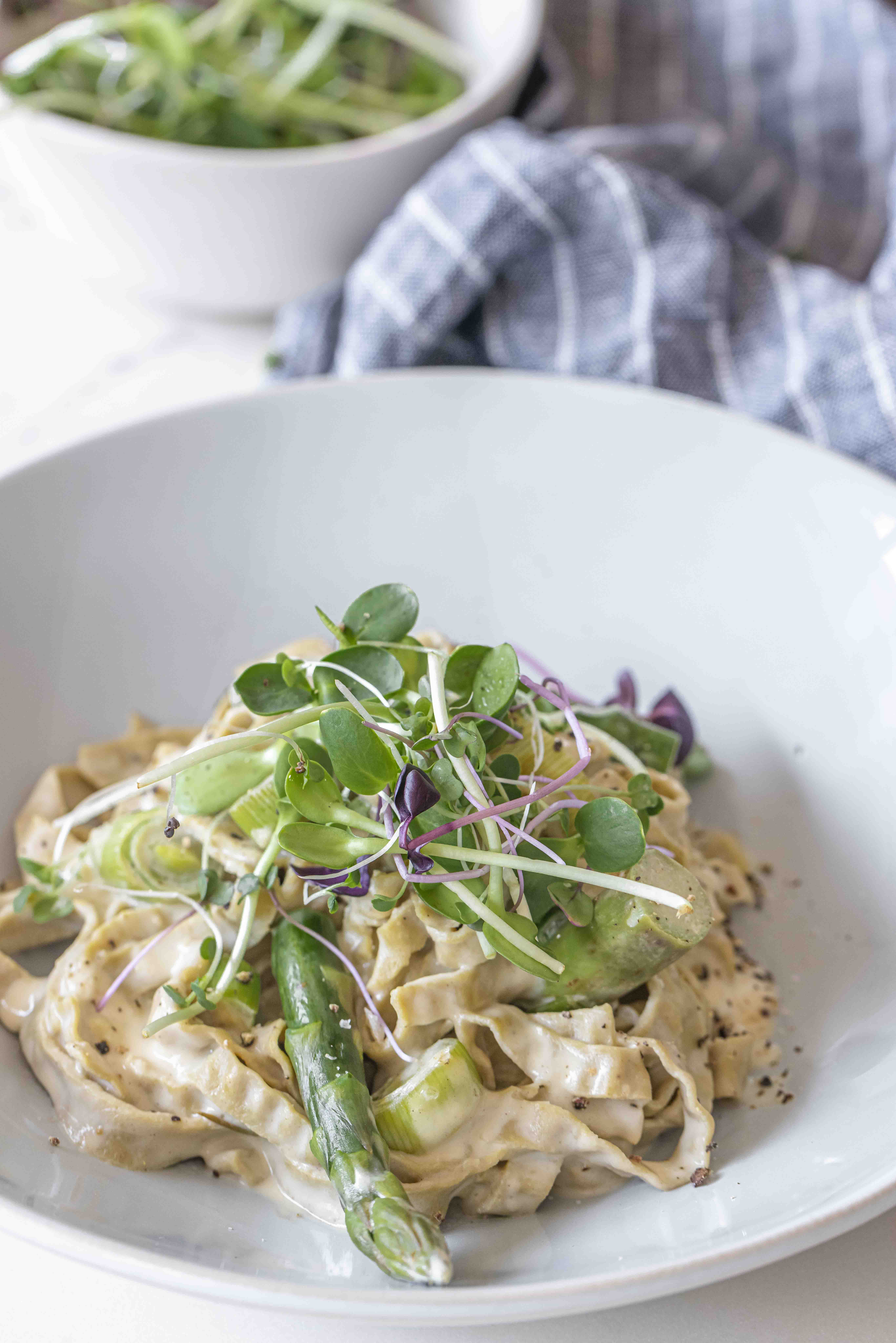 close up of Leek and Asparagus Fettuccine Alfredo on white pasta bowl, topped with micro greens around blue table cloth
