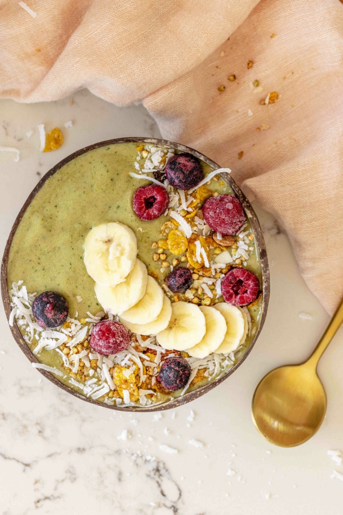 top down of matcha smoothie bowl with gold spoon, pink cloth and topped with berries, granola and banana