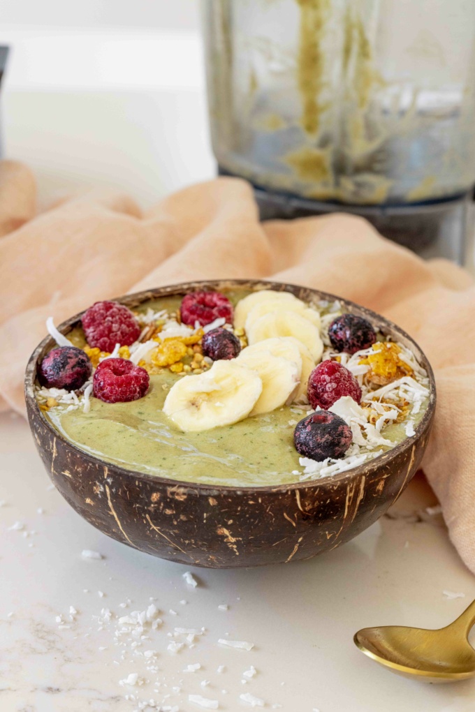 matcha smoothie bowl with gold spoon, pink cloth and topped with berries, granola and bananaa